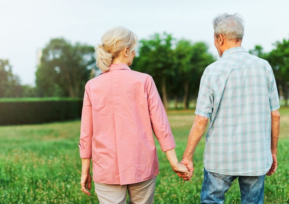 Elderly couple holding hands, walking across a grassy field. Trees in the background.