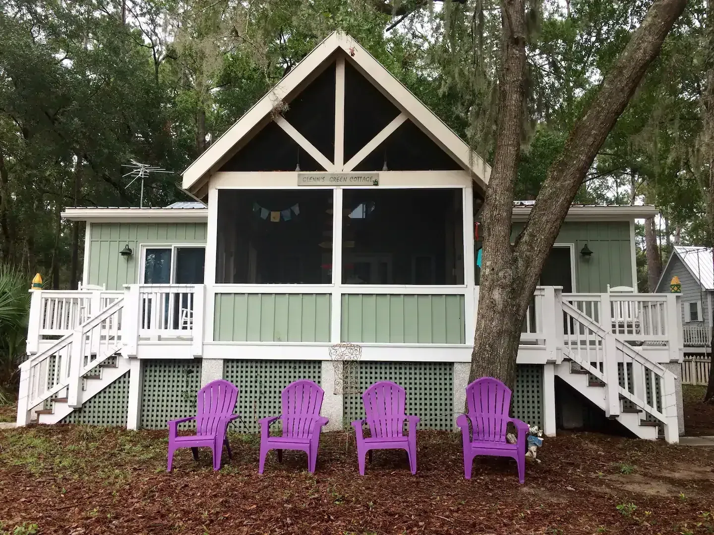 Cottage with a screened porch, purple chairs, and stairs, surrounded by trees.