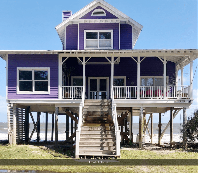 Purple beach house on stilts with porch and wooden stairs leading to the entrance, blue sky background.