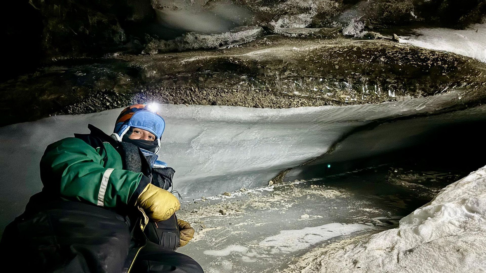 A man is kneeling in the snow in a cave with a headlamp.
