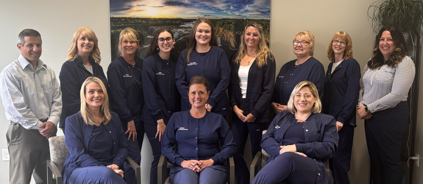 Group photo of dental office staff in navy scrubs, posing in front of a neutral wall with art.