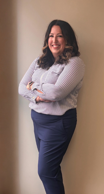 Woman with arms crossed, smiling, wearing striped shirt and blue pants, leaning against a wall.