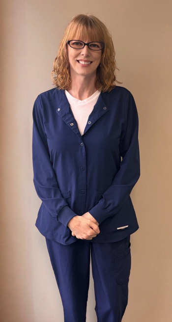 Woman with glasses smiles at the camera, light skin, brown hair, wearing a black shirt, seated indoors.