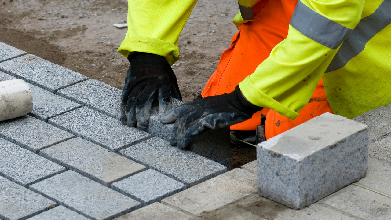 Construction worker placing paving stone; wearing neon yellow and orange safety gear.