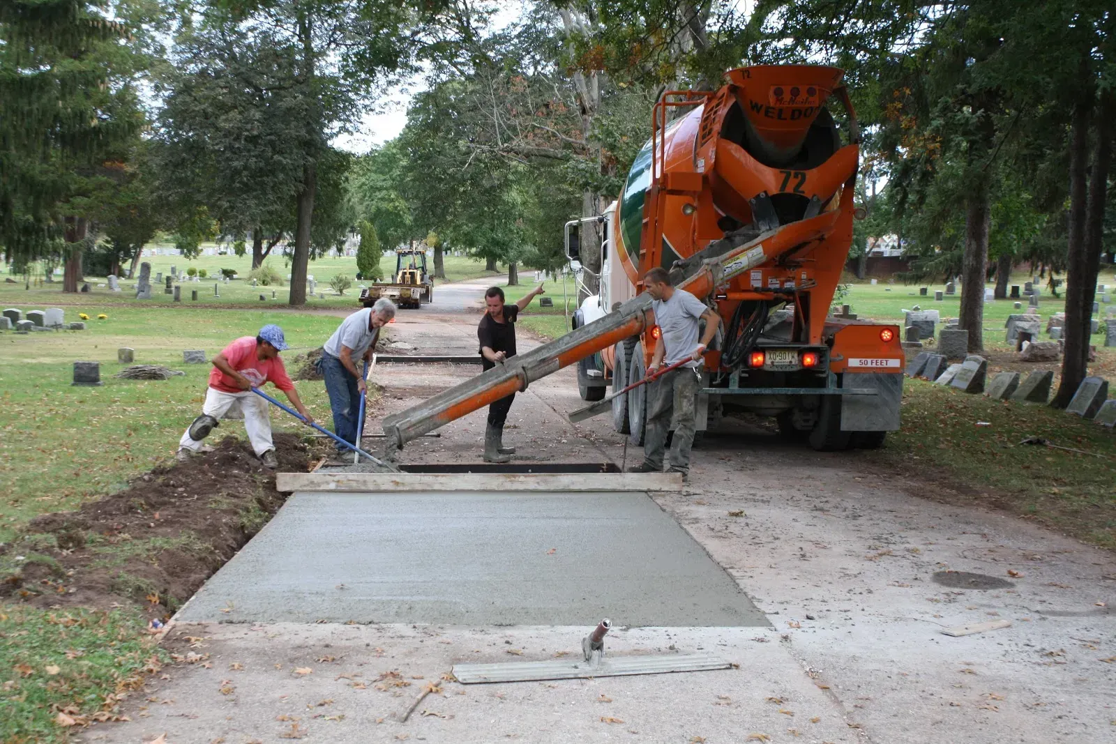 Cement truck pouring concrete onto a new pathway in a cemetery, workers smoothing the wet concrete.