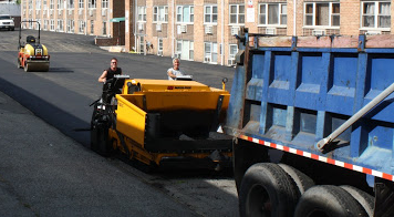 Road paving in progress. Workers operating machinery, asphalt laid, dump truck present.