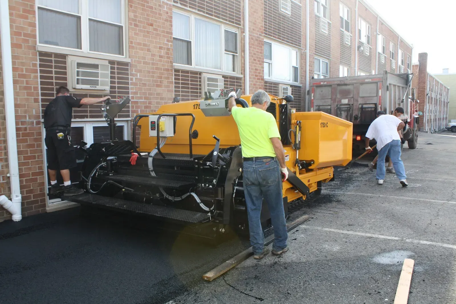Construction workers paving asphalt in a parking lot next to a brick building; yellow machinery.