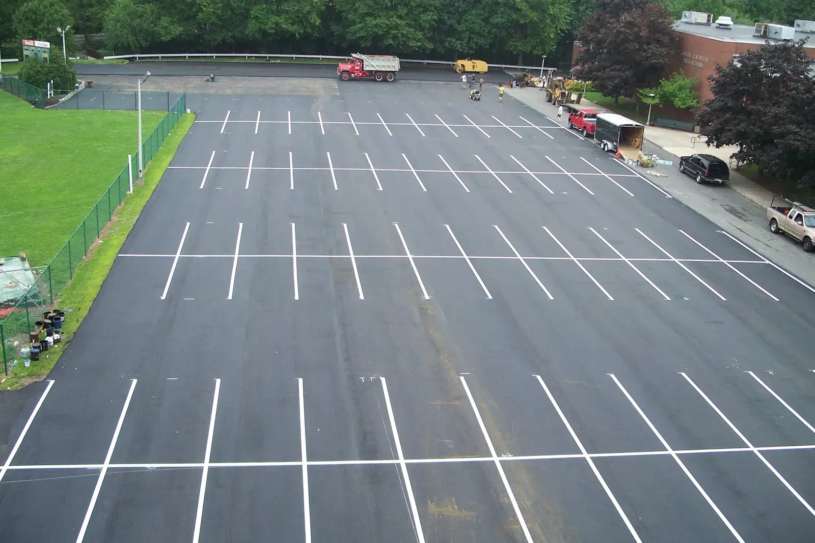 Empty parking lot with freshly painted white lines. Trucks and building visible. Green grass and trees nearby.