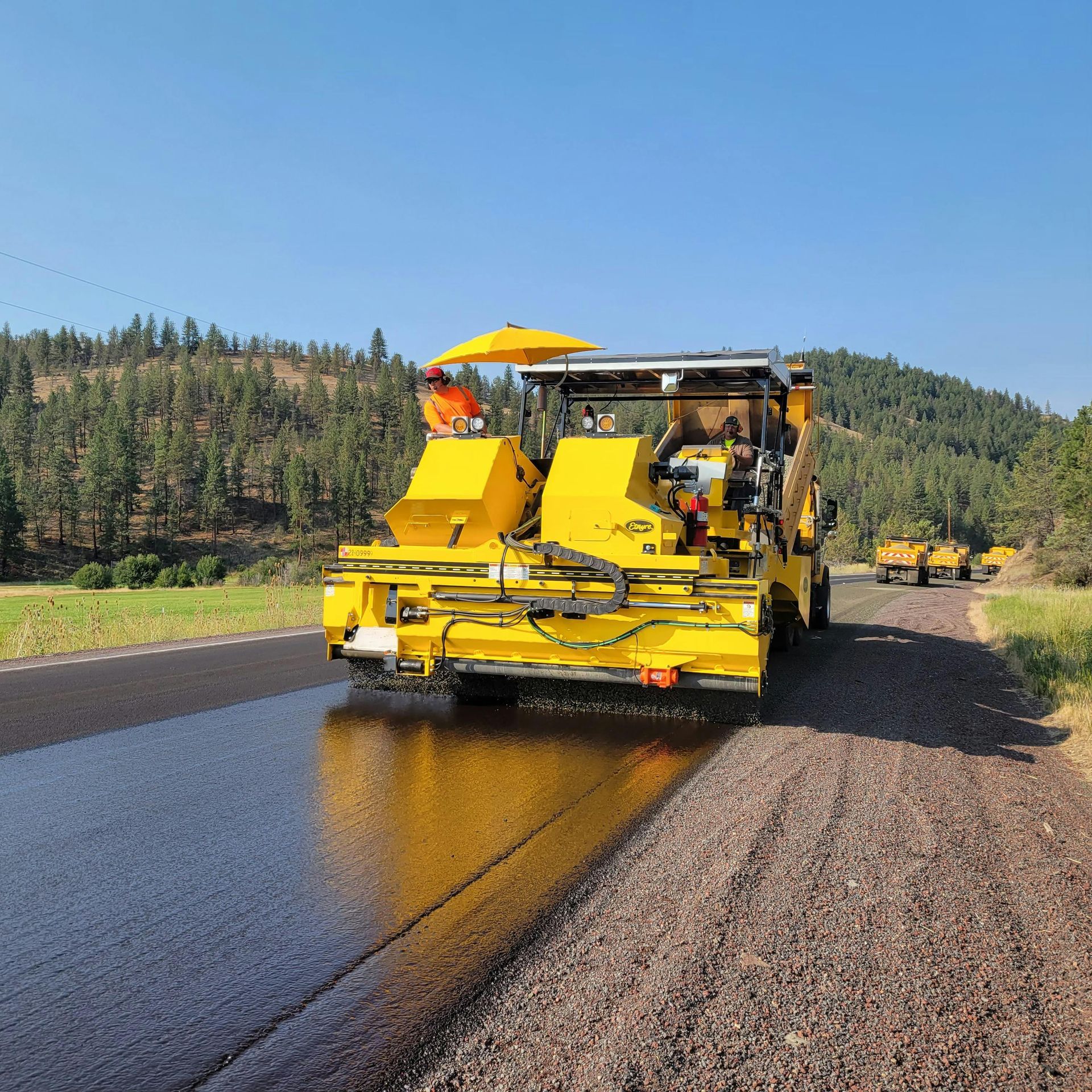 A bright yellow asphalt paver moves along a road, applying a dark liquid sealant over a rocky, unfinished surface.