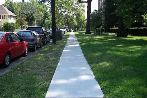 Sidewalk along parked cars and grassy area with trees under a sunny sky.