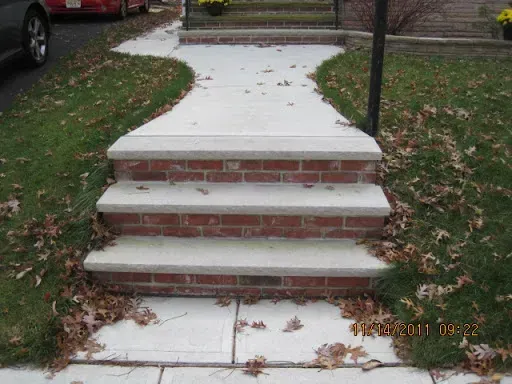 Brick steps leading to a concrete walkway, bordered by green grass and fallen leaves.