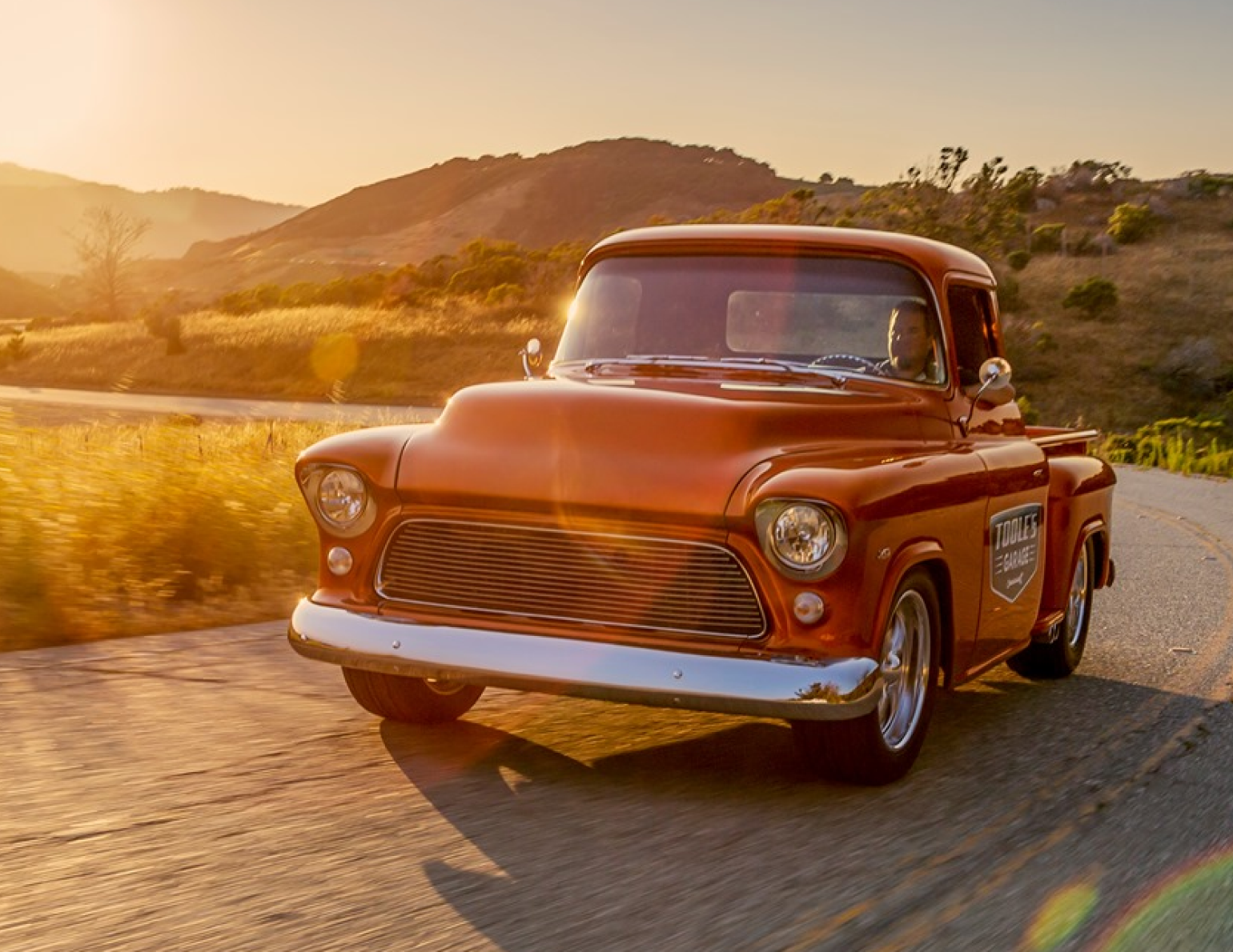 An old orange truck is driving down a country road at sunset.