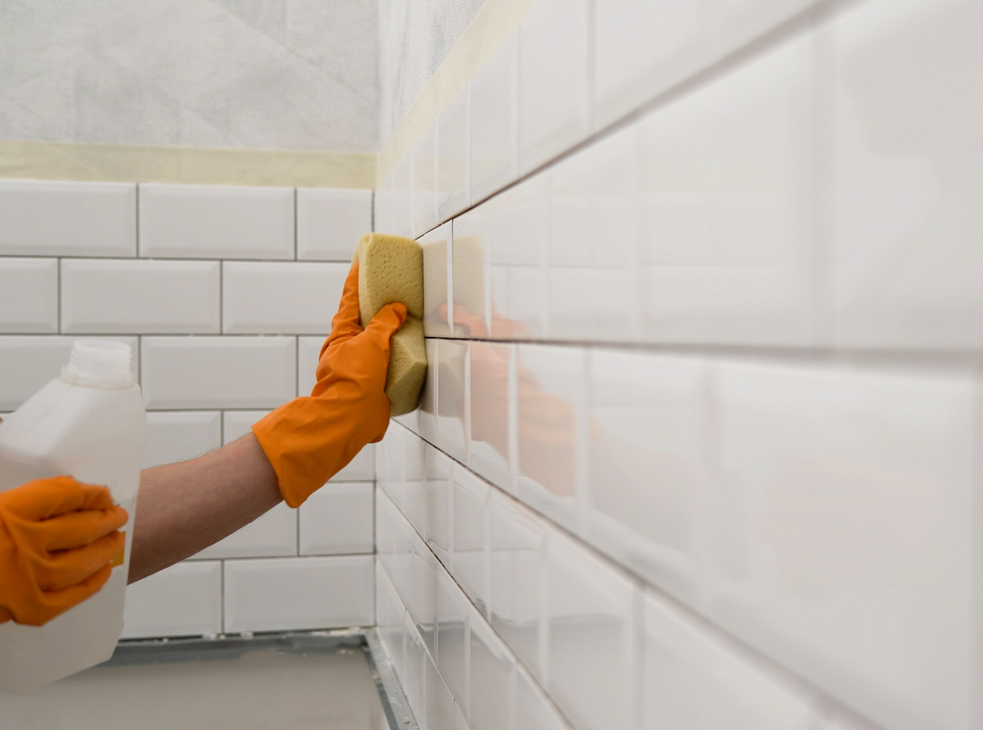 A person wearing orange gloves is cleaning a white tile wall with a sponge.