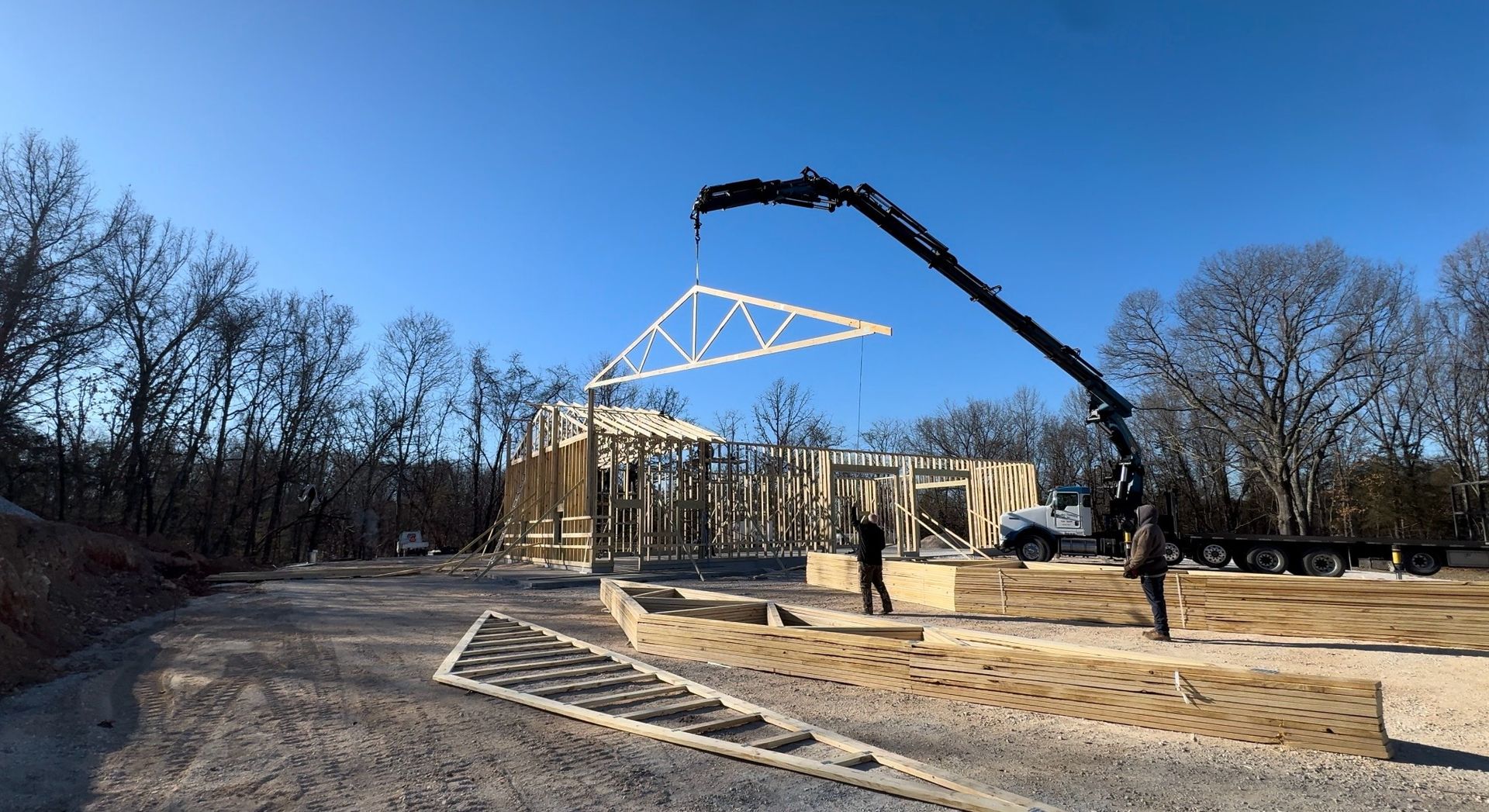 A crane lifts a wooden roof truss into position over a structure under construction in a wooded, sunny area.