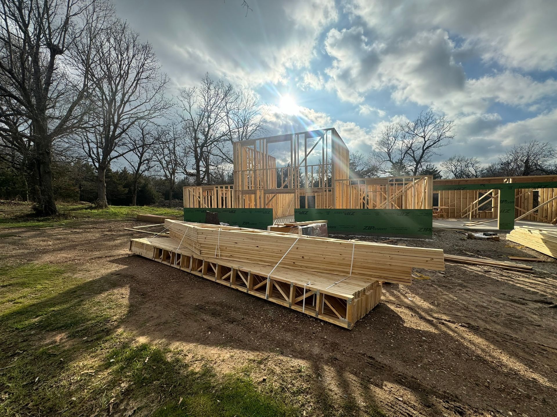 A wooden house frame under construction in a grassy field with a stack of floor trusses in the foreground.