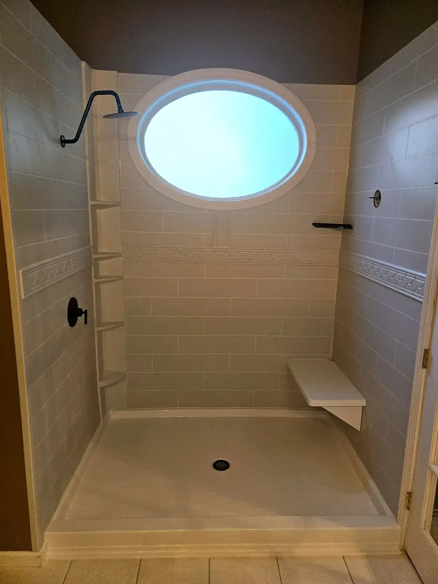 Shower with gray tile, round window, black shower fixtures, and a built-in seat.