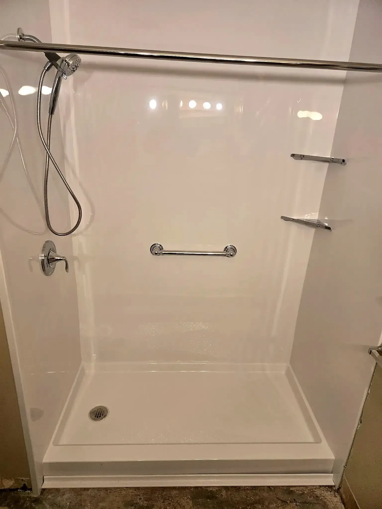 A white, walk-in shower stall featuring a handheld shower head, a horizontal grab bar, and two corner storage shelves.