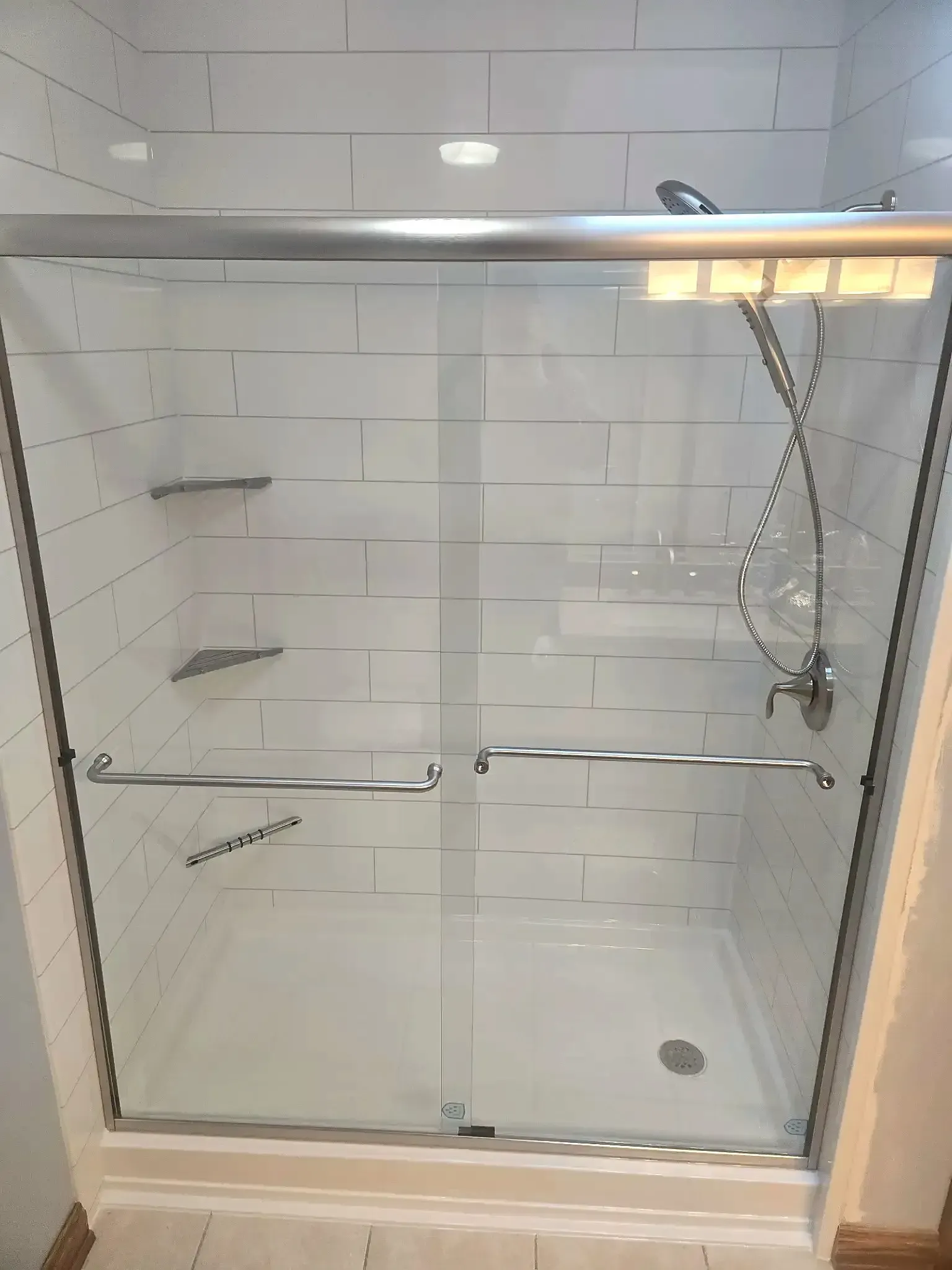 A walk-in shower with sliding glass doors, white subway tile walls, built-in corner shelves, and a handheld shower head.