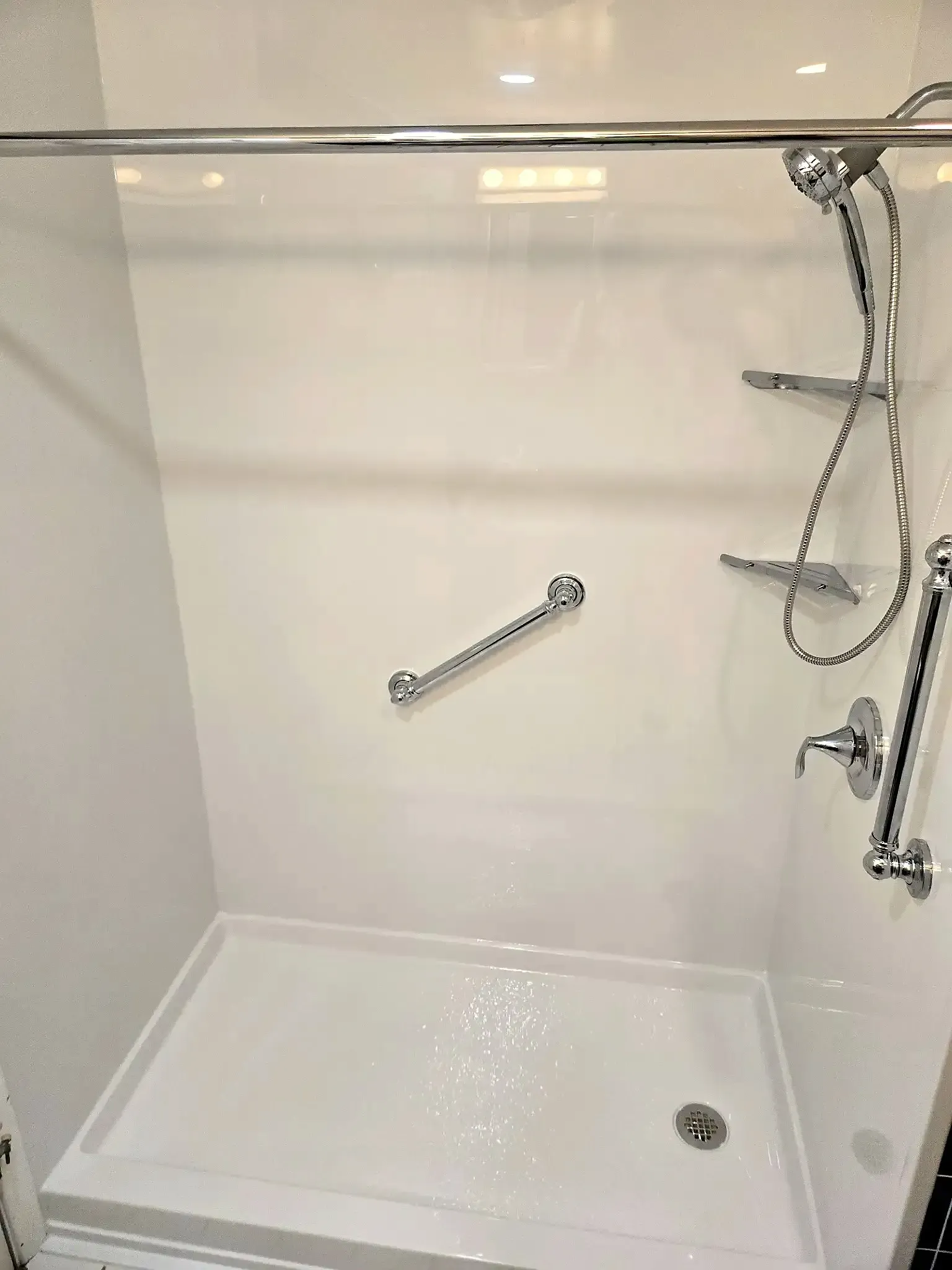A white walk-in shower stall with chrome fixtures, a handheld showerhead, two corner shelves, and a diagonal grab bar.