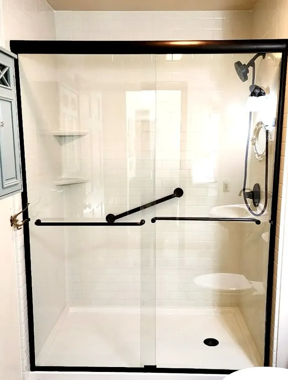 A shower stall with clear glass sliding doors, a black frame, black handles, a grab bar, and built-in white corner shelves.