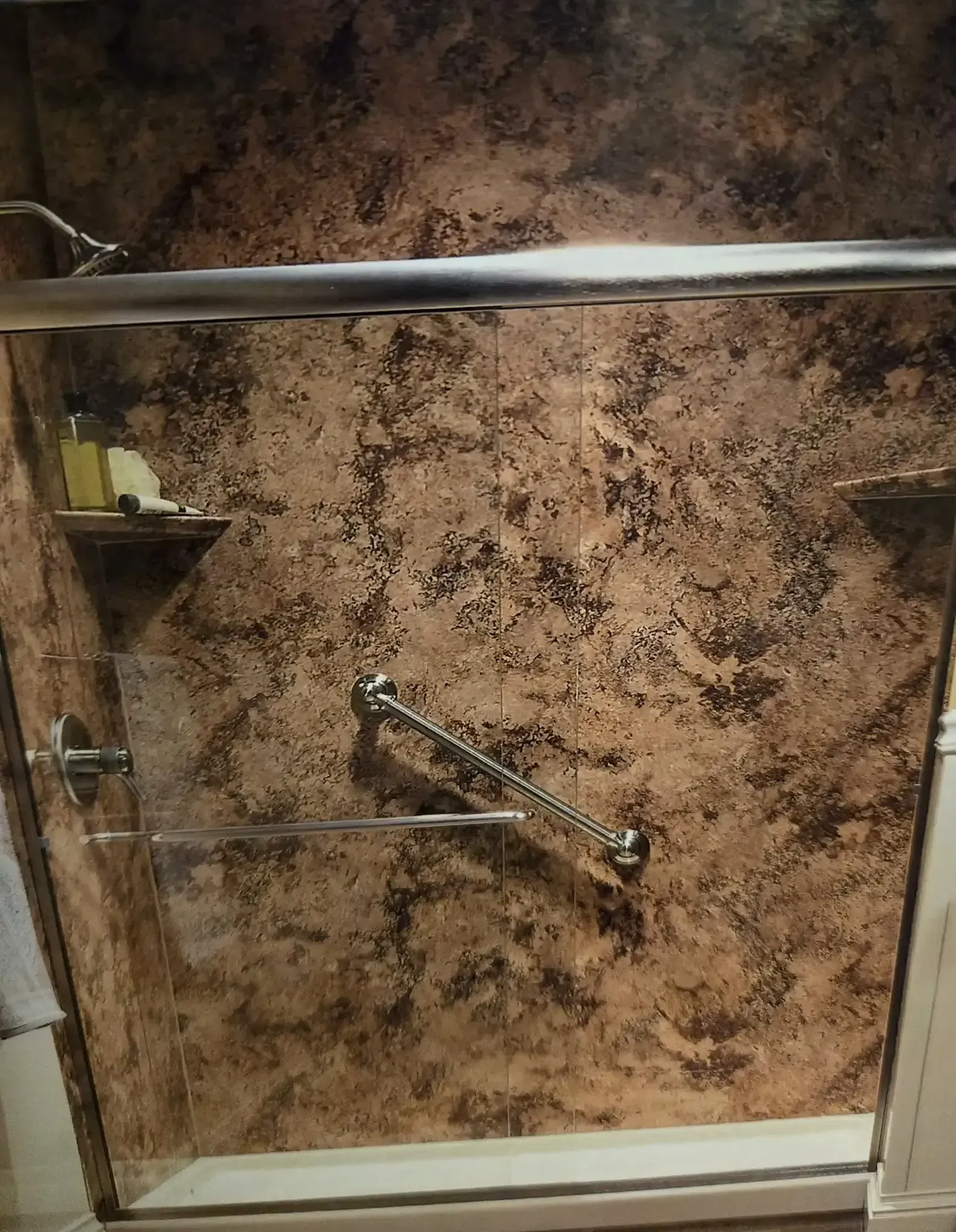 A shower stall with textured, mottled brown walls, a chrome grab bar, a glass door, and a built-in corner shelf.