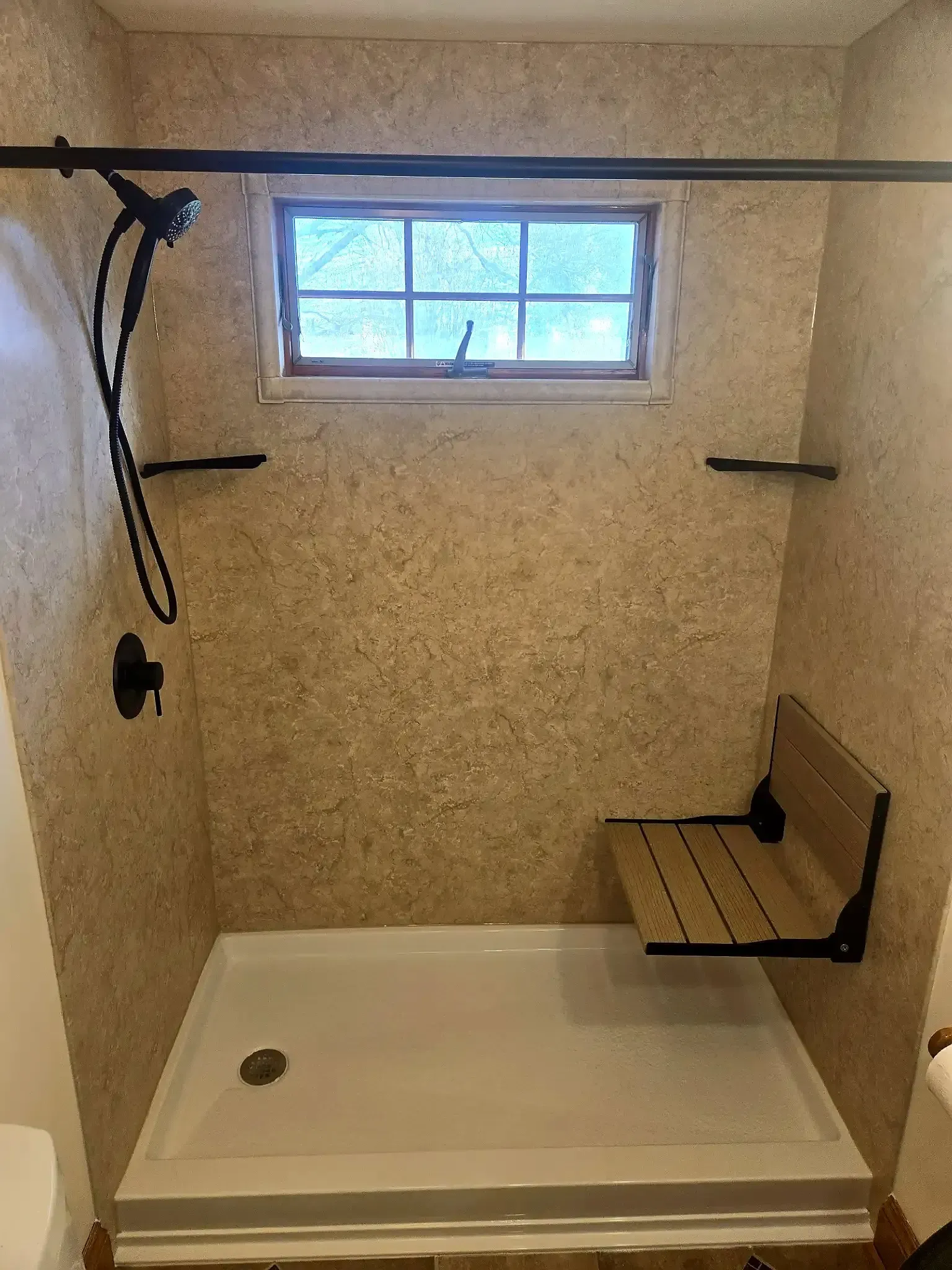 A walk-in shower with beige textured walls, a white floor, a black handheld showerhead, and a mounted wooden shower seat.