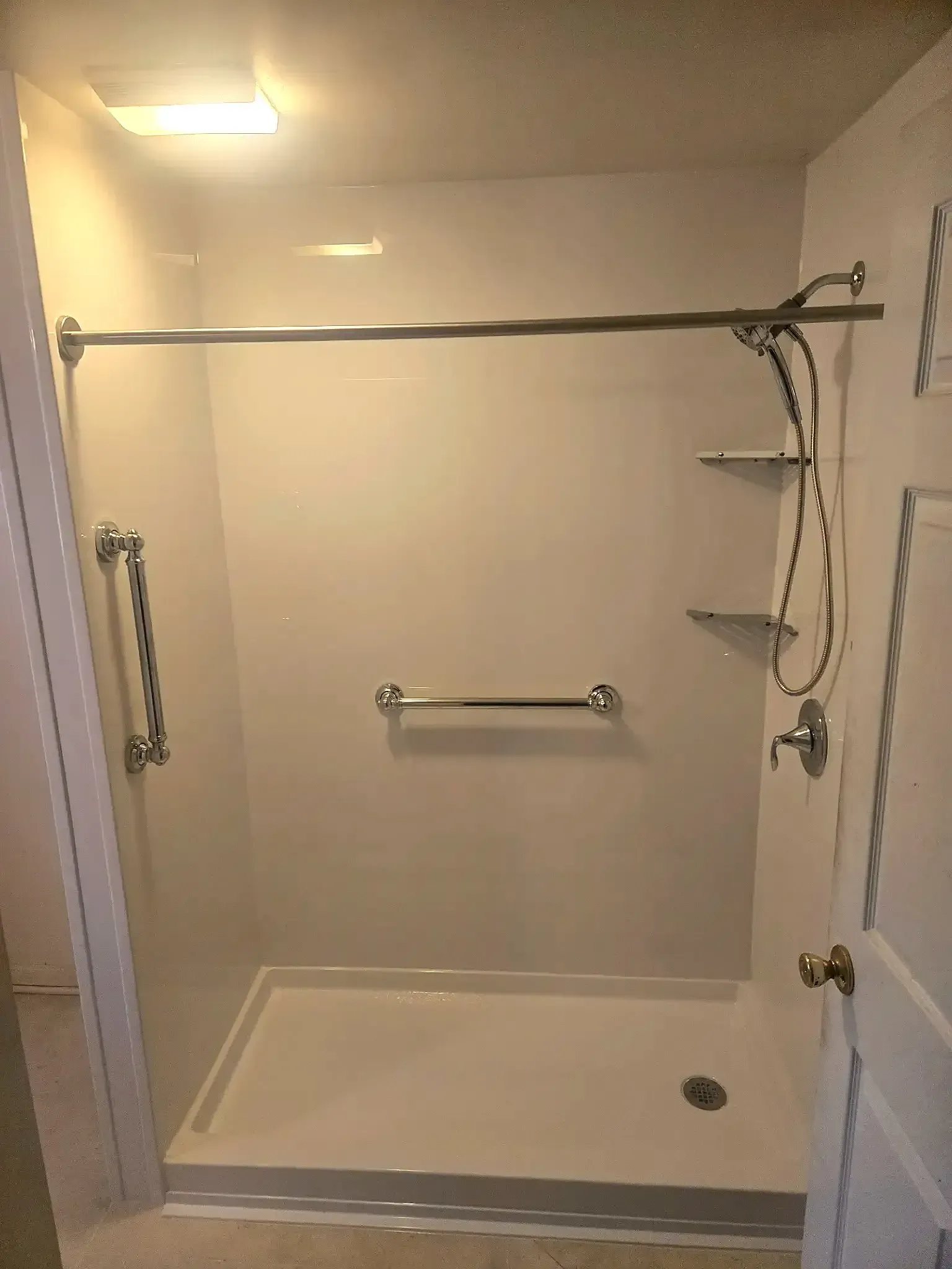 A white walk-in shower with grab bars on the side and back walls, integrated shelving, and a metal shower rod.
