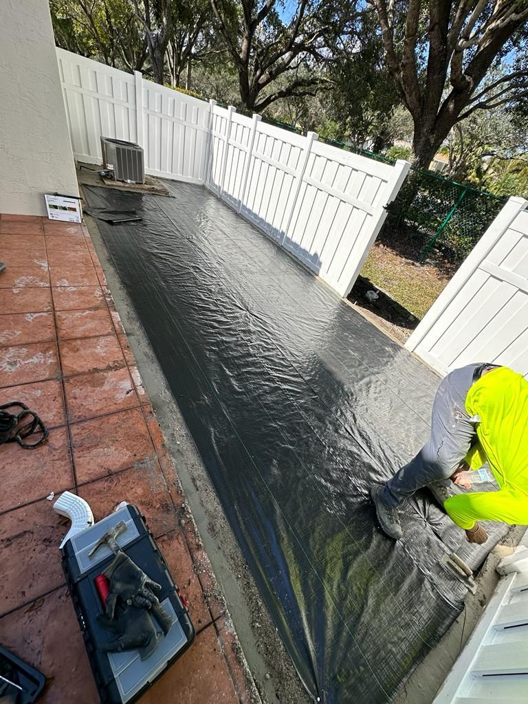 A man is laying concrete on a patio next to a white fence.