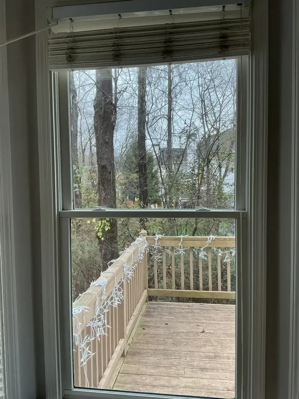 View through a window to a wooden deck and railing with trees beyond, framed by white blinds.
