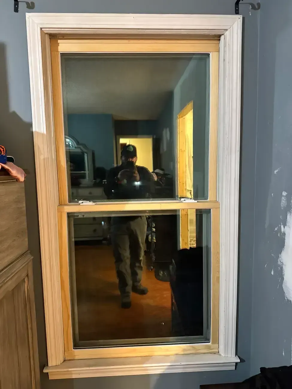 Small wooden-framed window set in a gray wall, reflecting a person standing inside a dim room.