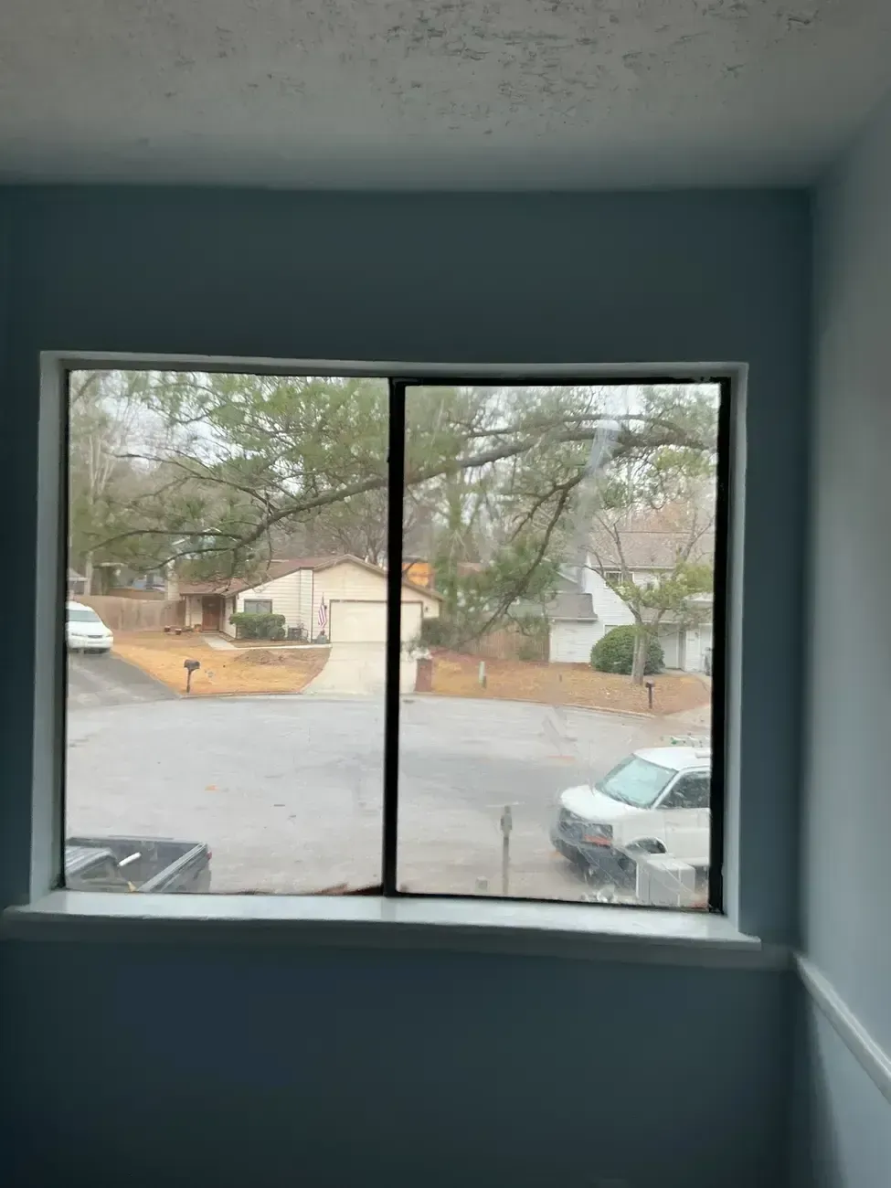 View through a window of a quiet residential street with parked cars and trees on a rainy day