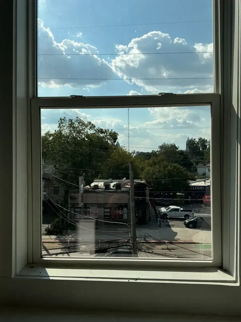 View through a window of trees, parked cars, and a bright blue sky with clouds.