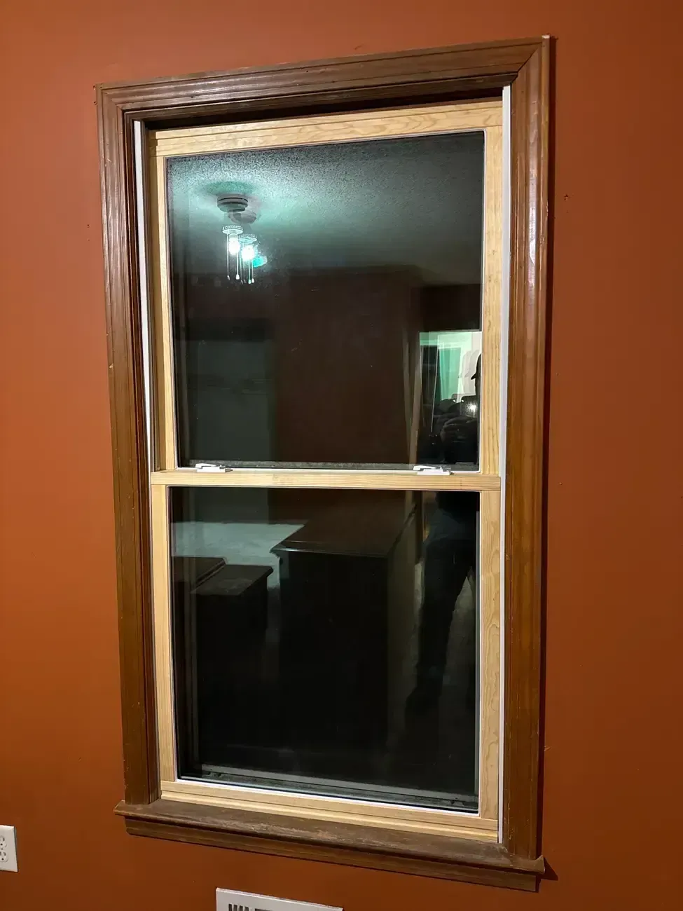 Wood-framed wall mirror reflecting a dim room and ceiling light on a brown wall