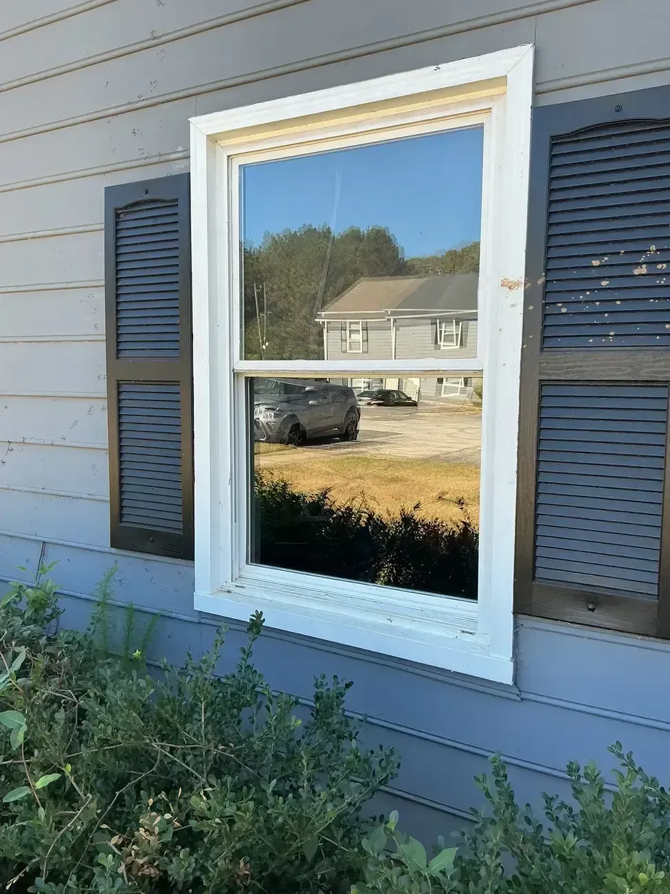 White-trimmed window with dark shutters on a gray house exterior, reflecting a yard and house across the street