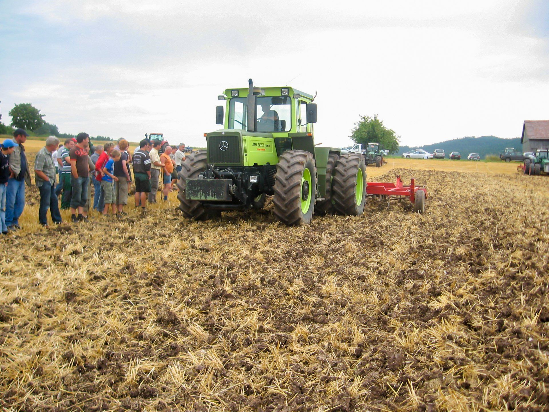 Ein grüner Traktor pflügt ein Feld, während eine Gruppe von Leuten zusieht.