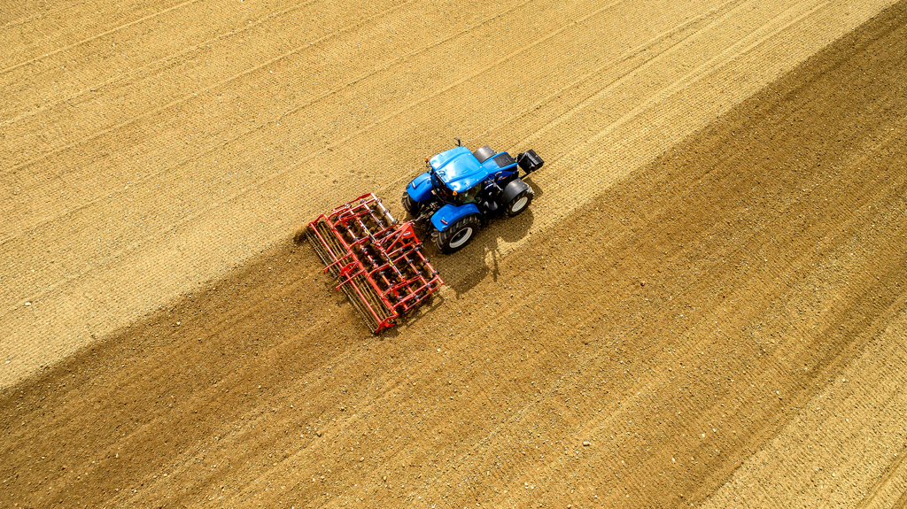 Ein blauer Traktor pflügt ein Feld und erzeugt kontrastierende Streifen im braunen Boden.