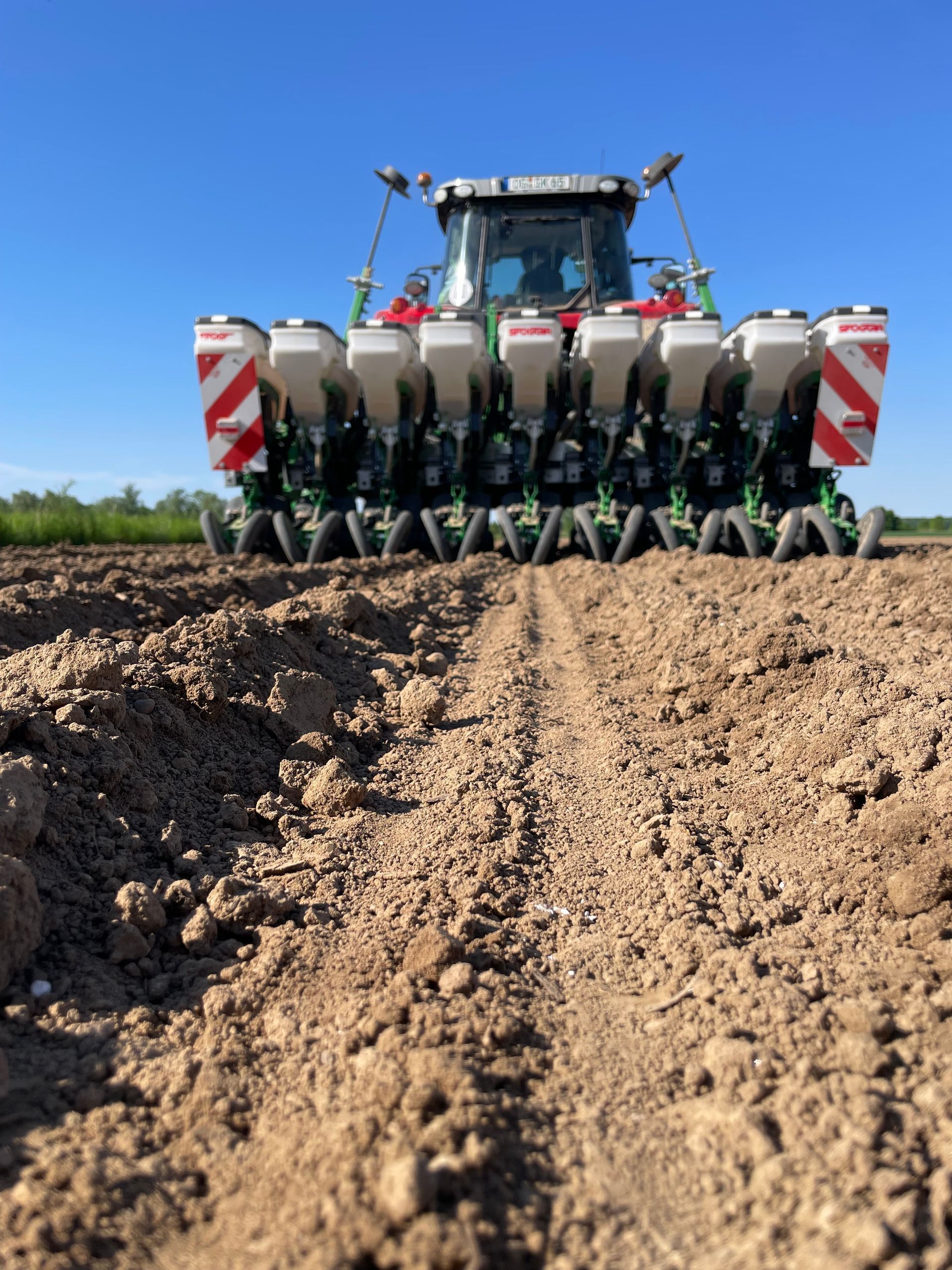 Ein Traktor sät Samen auf einem Feld. Die Maschine ist grün-weiß, der Himmel blau. Furchen sind im Boden zu erkennen.