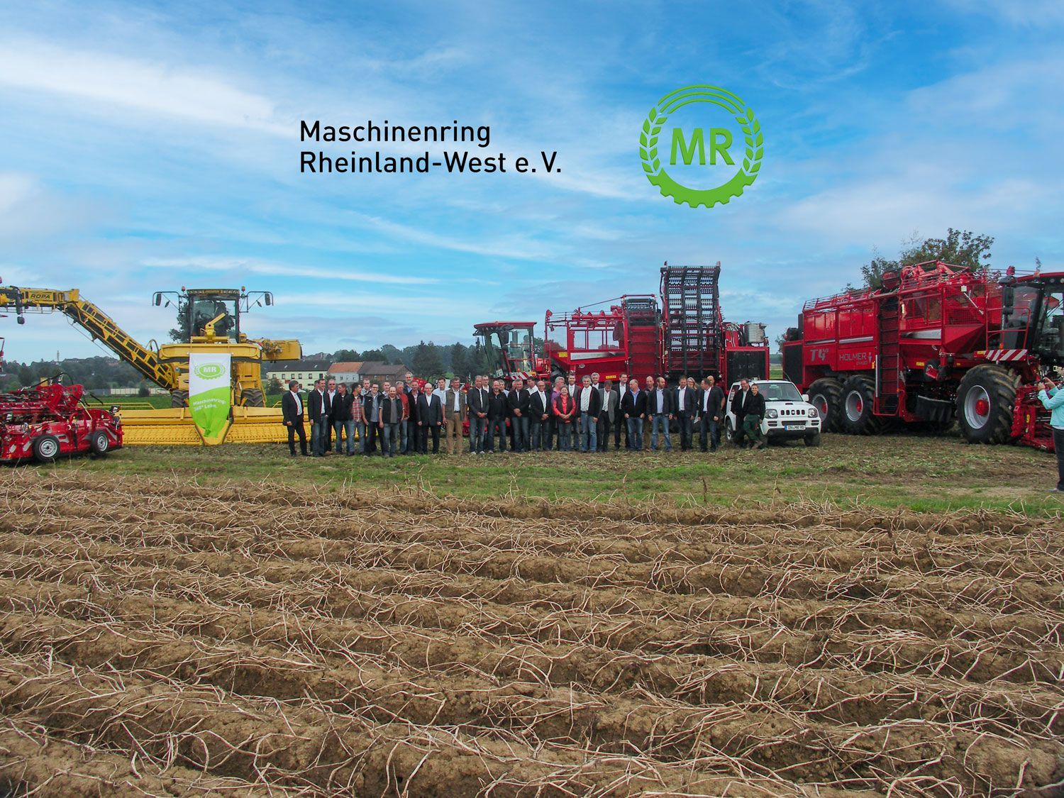 Gruppe von Menschen posiert mit landwirtschaftlichen Maschinen auf einem Feld; Logo sichtbar.