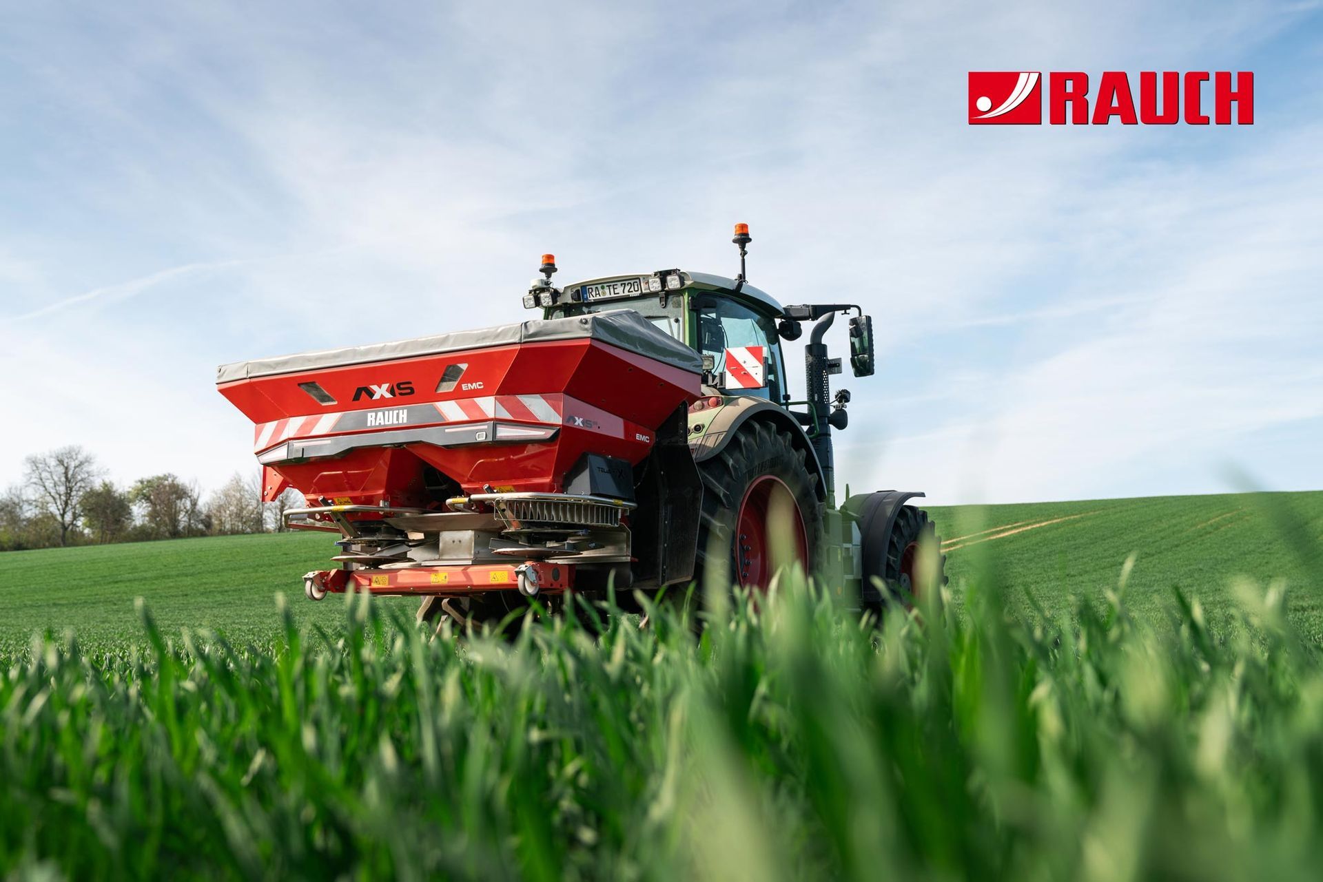 Ein roter Traktor streut Dünger auf ein grünes Feld unter blauem Himmel. Rauch-Logo in der Ecke.