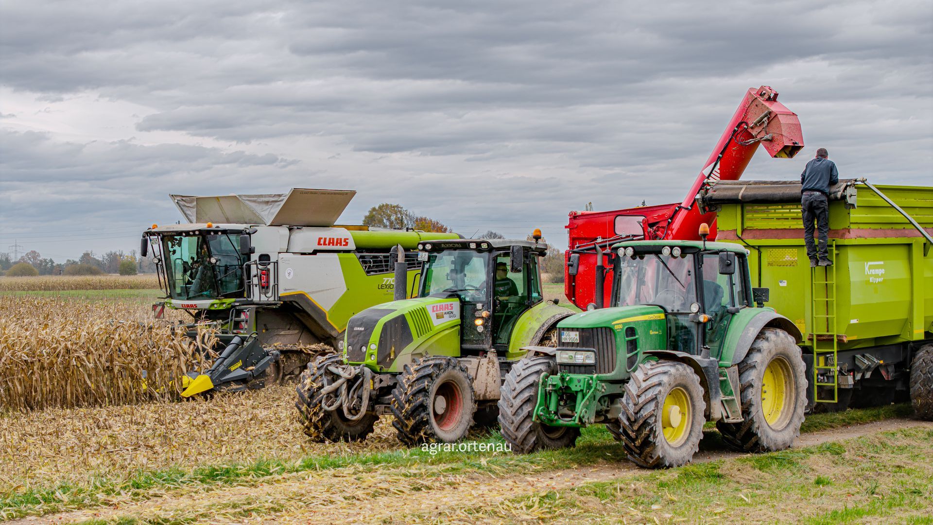 Maisernte: Mähdrescher, zwei Traktoren und ein Anhänger auf einem Feld unter bewölktem Himmel.