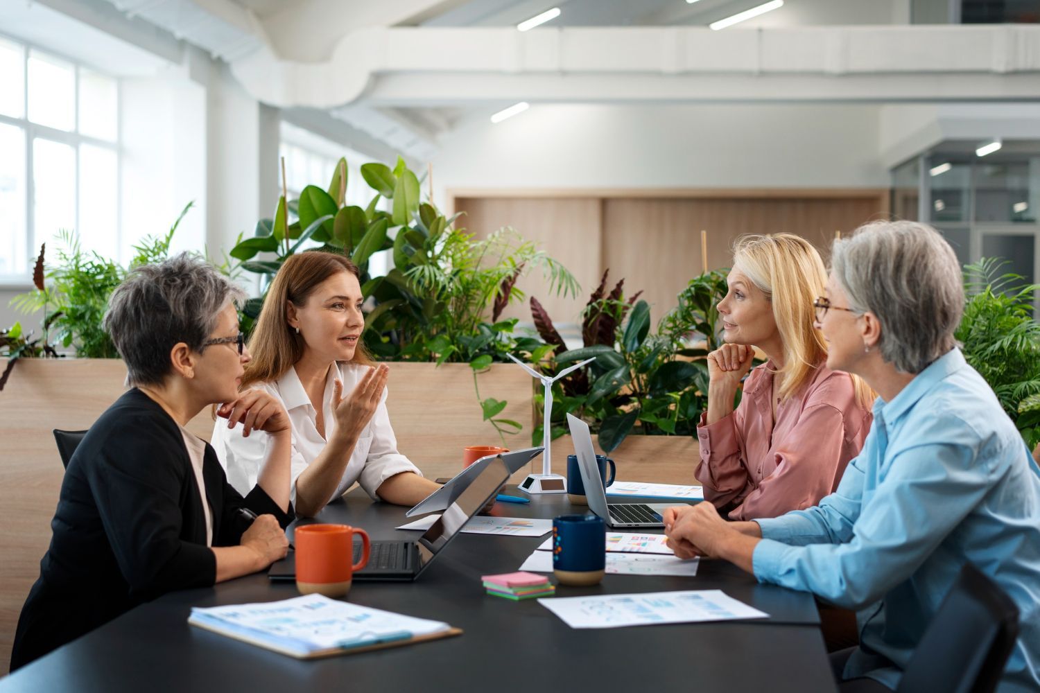 Four women in a modern office meeting, discussing at a table with laptops, plants, and documents.