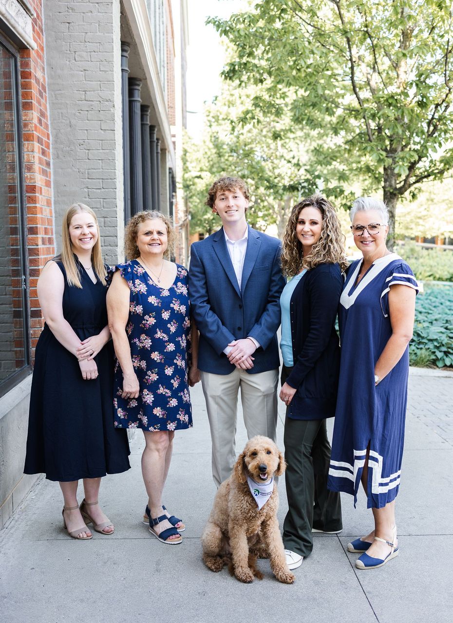 Group of six people and a dog posing in an office setting. Two seated men in blue blazers, others standing.