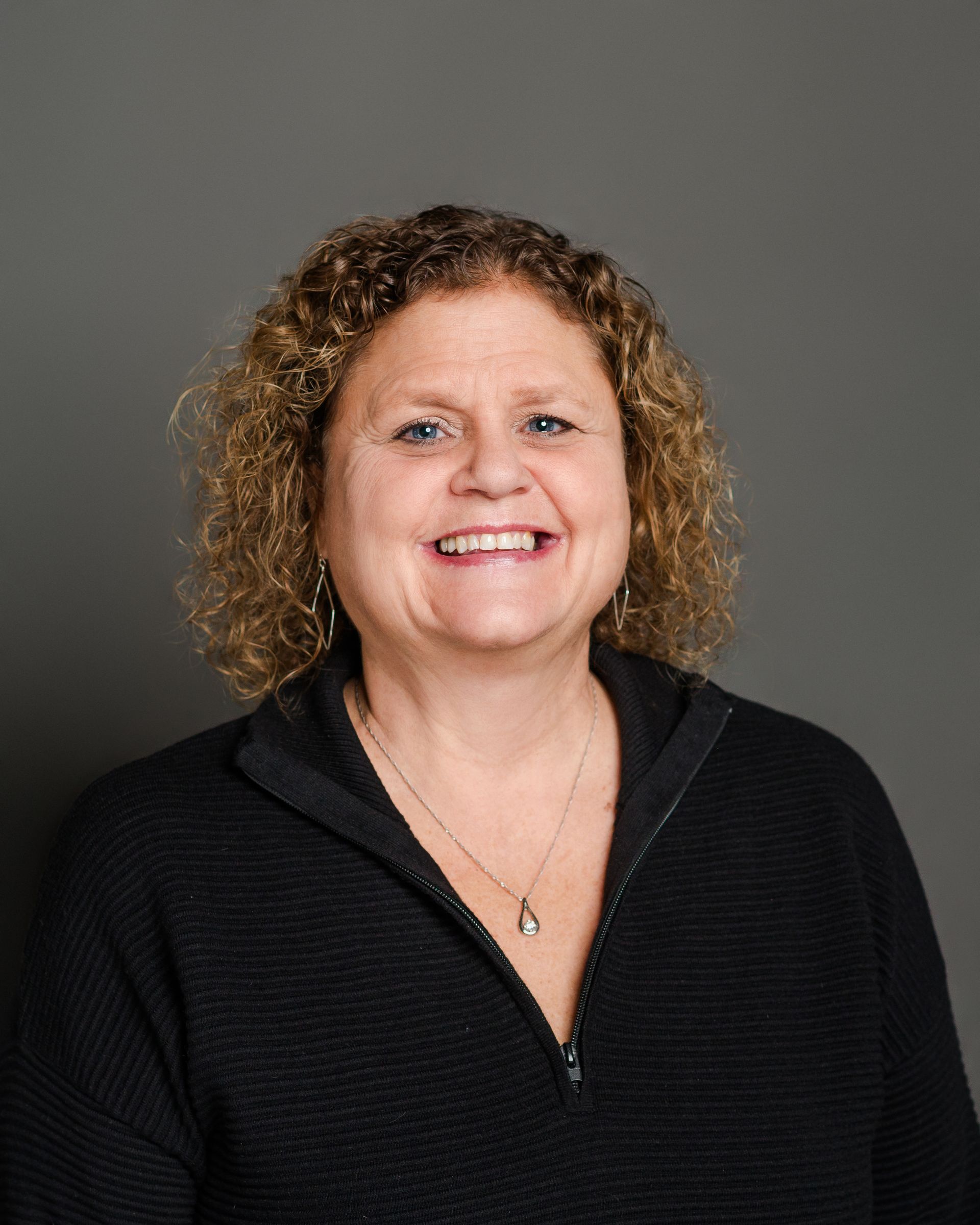 Woman with curly hair smiles, wearing a black zip-up sweater, gray background.
