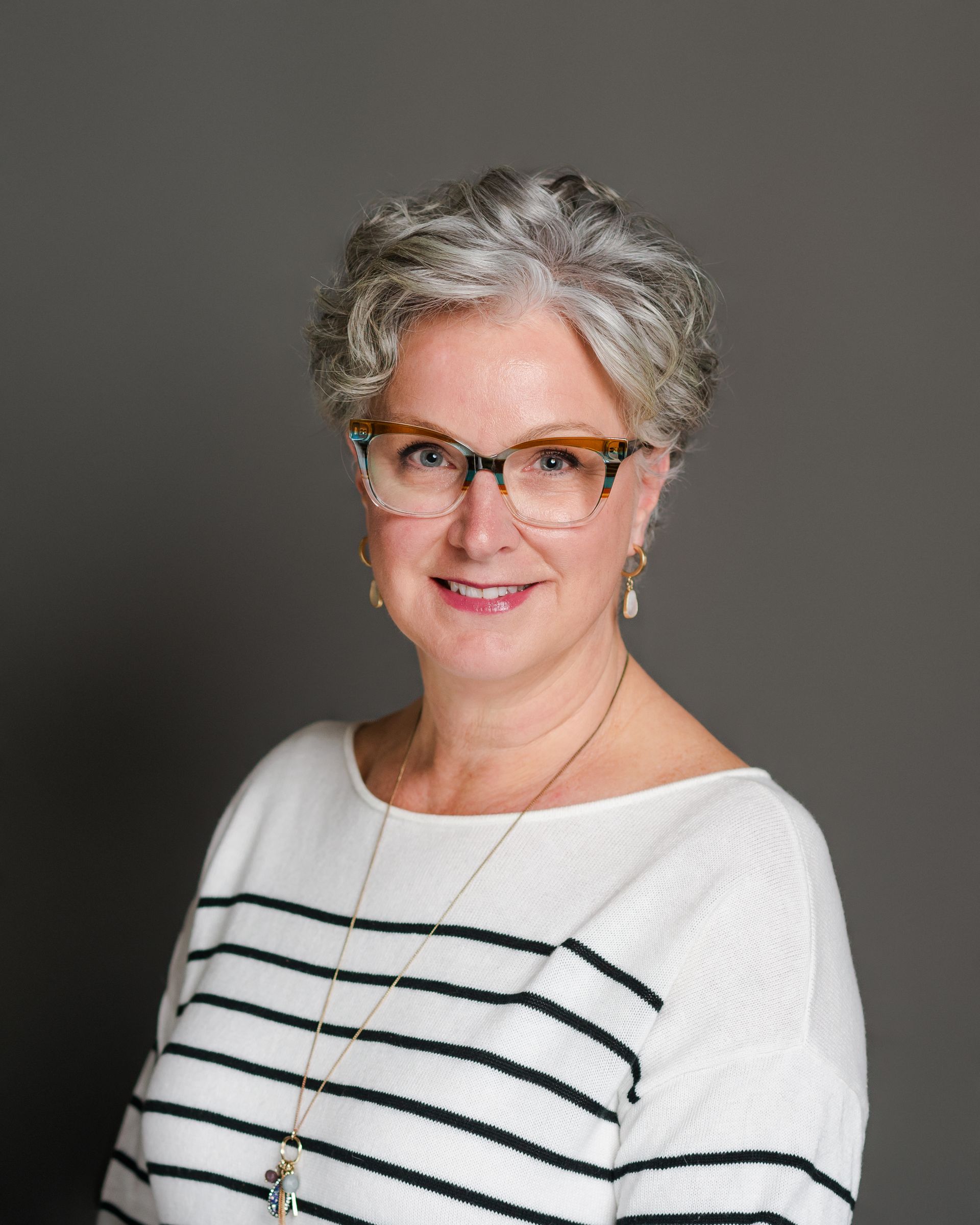 Woman with short gray hair and glasses smiles. Wearing a striped shirt and necklace, standing against a gray backdrop.