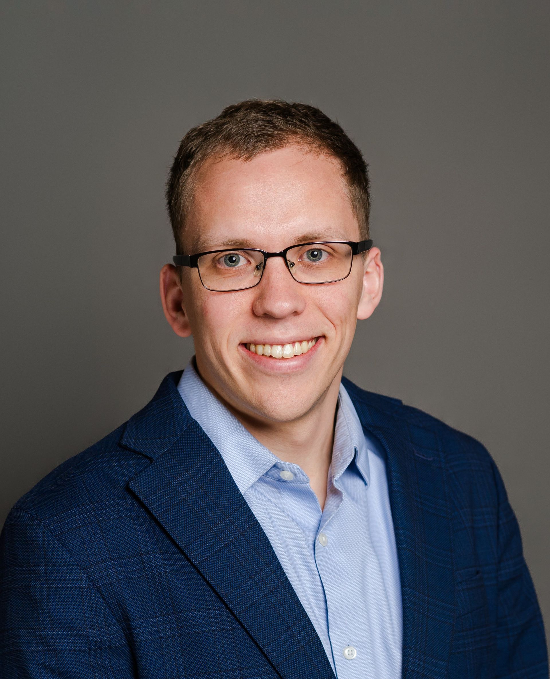 Man wearing glasses and blue blazer smiles at the camera against a grey background.