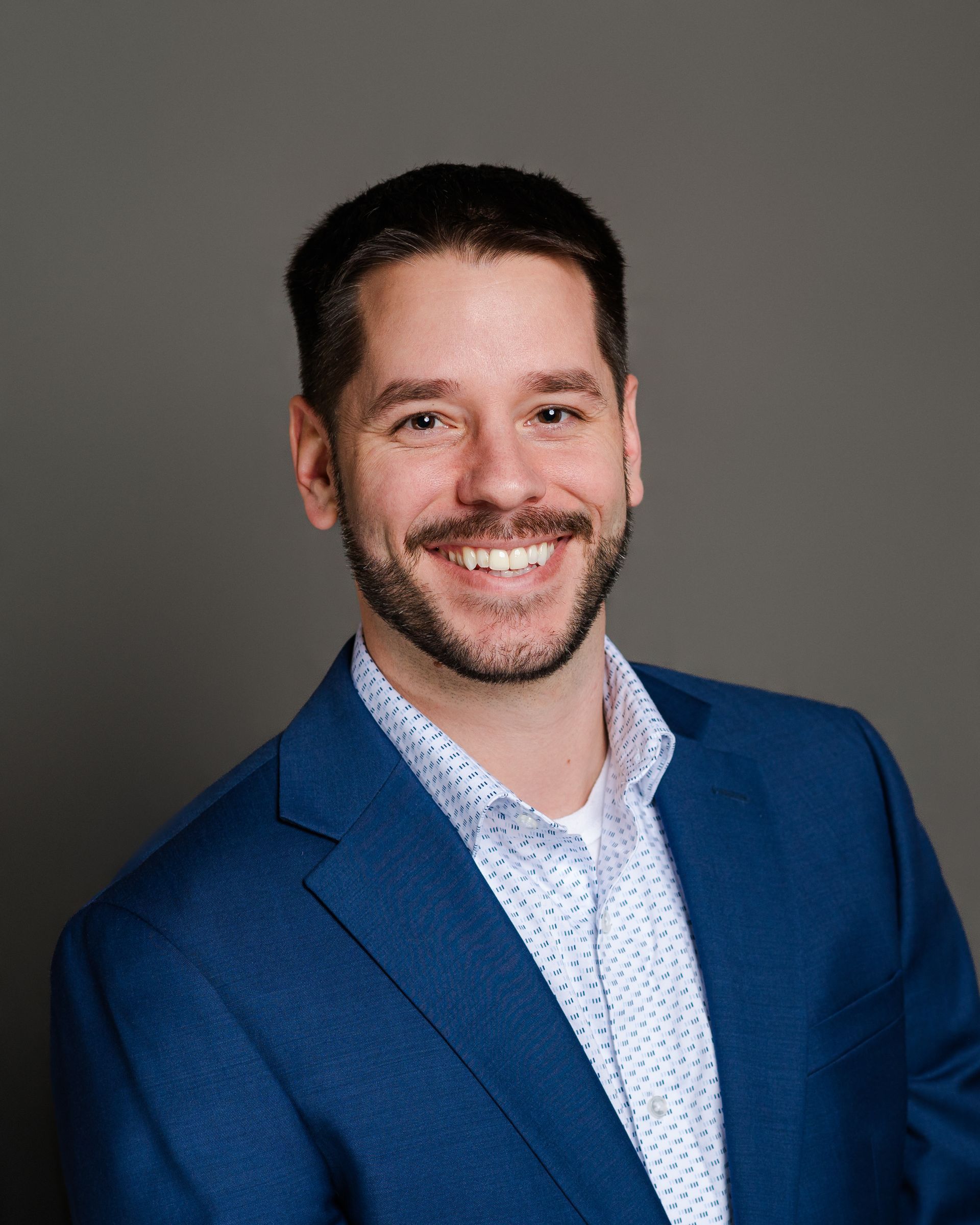 Man in blue blazer smiling, looking at the camera against a grey backdrop.