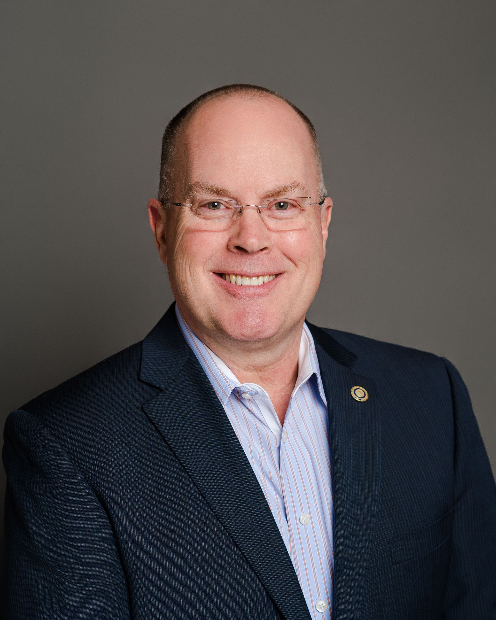 Smiling man in glasses, wearing a blue blazer and light blue shirt, against a gray background.