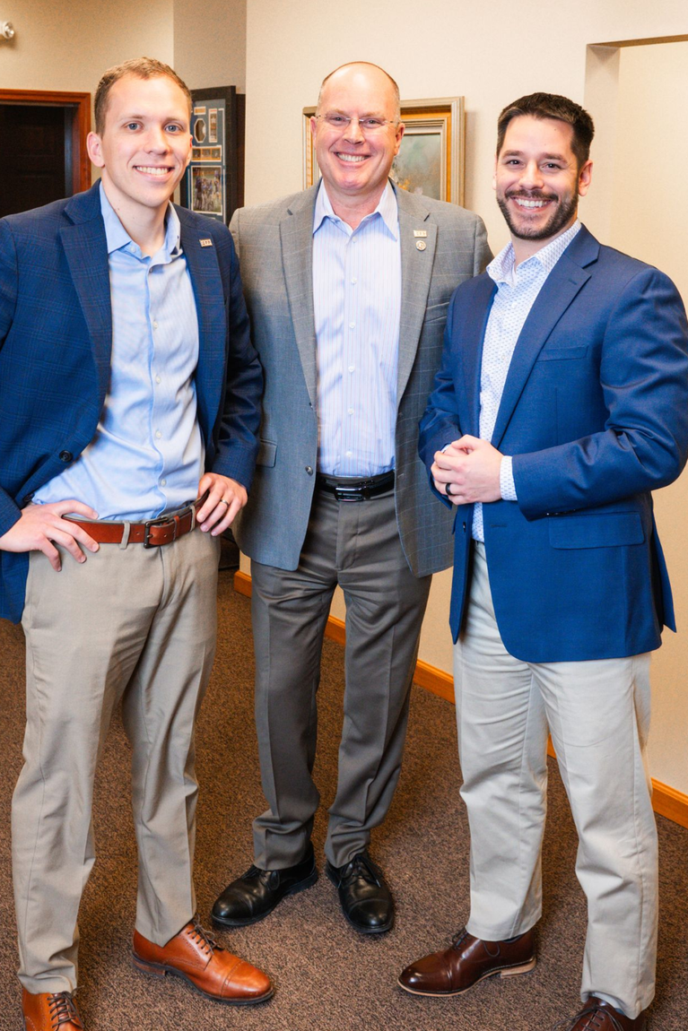 Three men in business attire standing indoors, smiling.