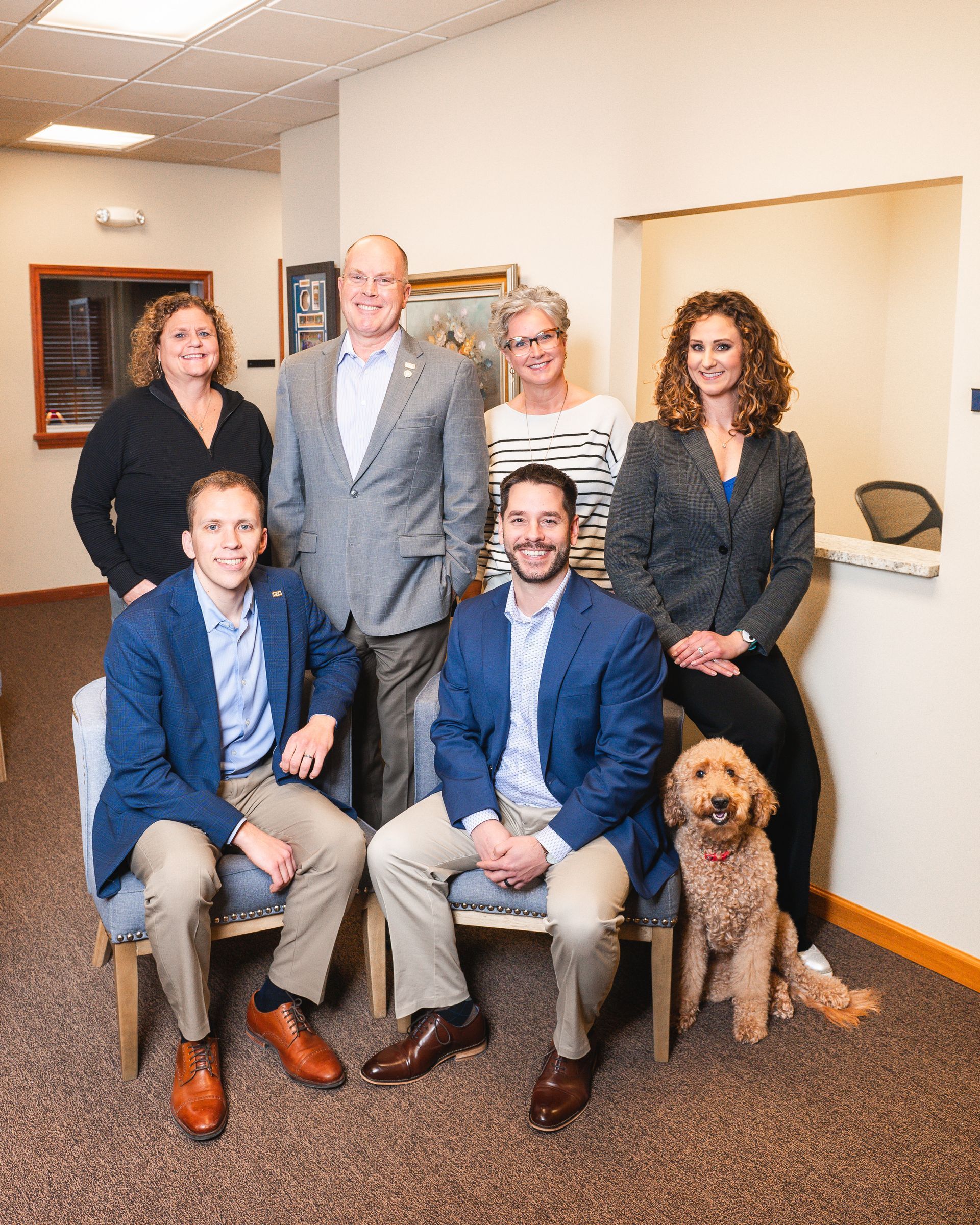 Group of six people and a dog posing in an office setting. Two seated men in blue blazers, others standing.