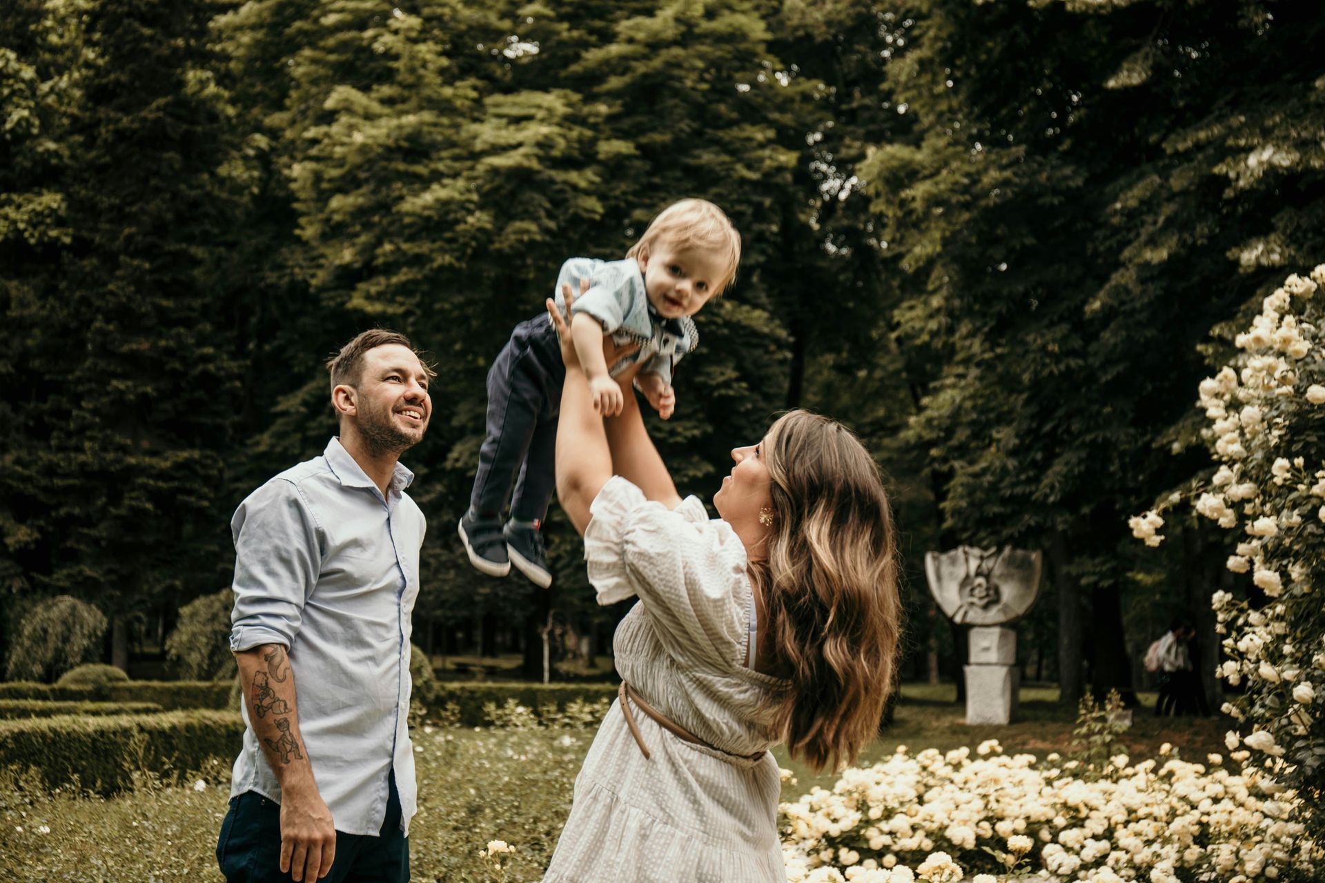 Family in a park: mother tossing toddler up in the air, father watches smiling, surrounded by flowers and greenery.